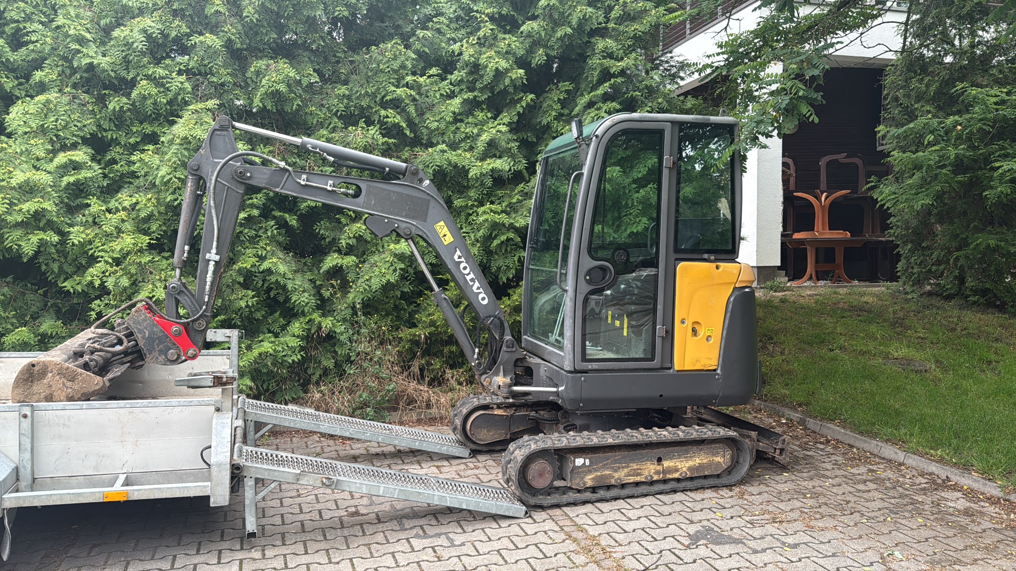 Szara minikoparka Volvo na przyczepie z najazdami, z łyżką załadowaną ziemią, na tle zieleni i budynku. Sprzęt gotowy do transportu.