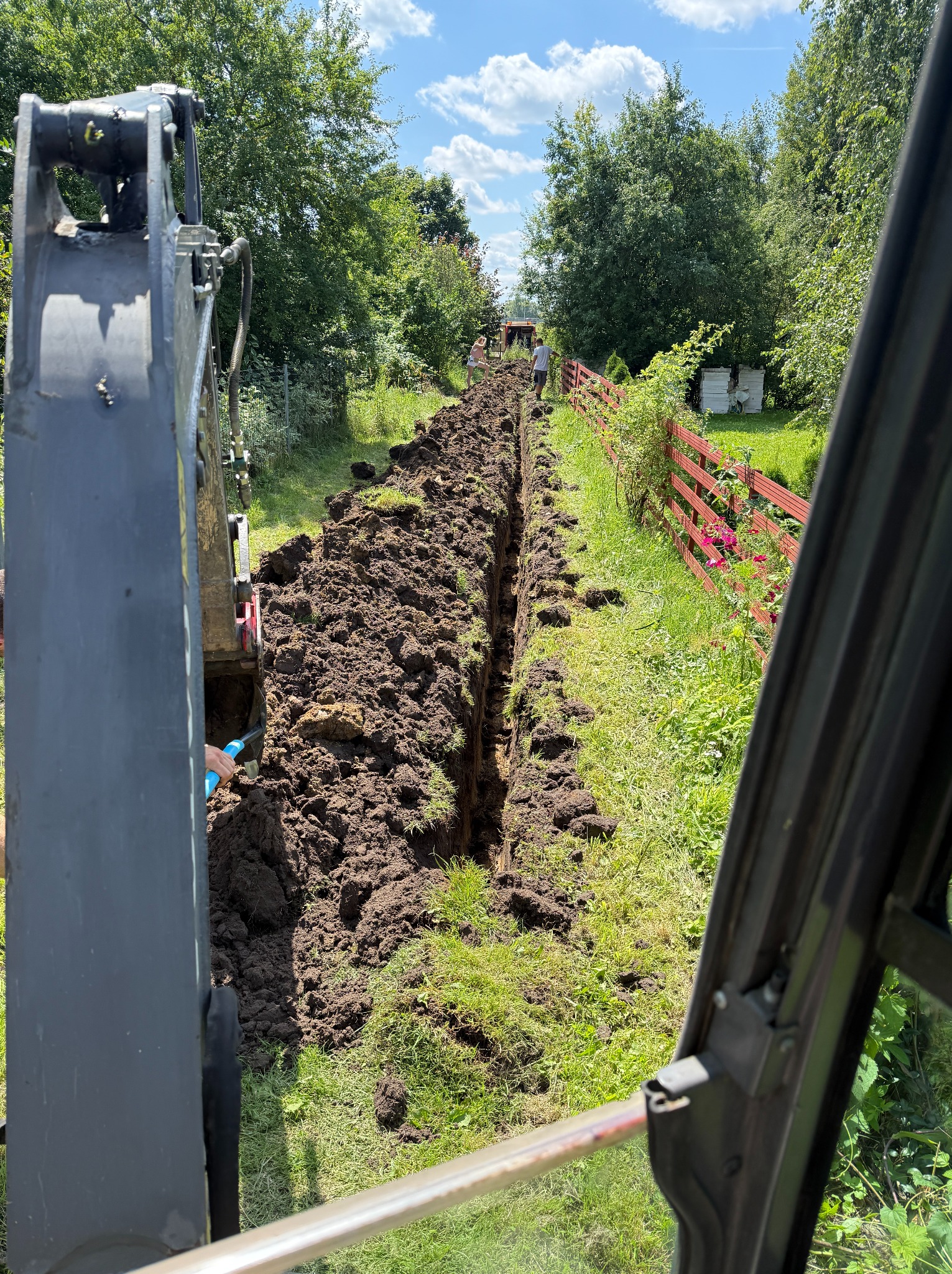 Wykopany wąski rów w ziemi, widok z kabiny koparki, z boku czerwony płot i zieleń. W rowie widoczna niebieska rura. Prace ziemne w słoneczny dzień.