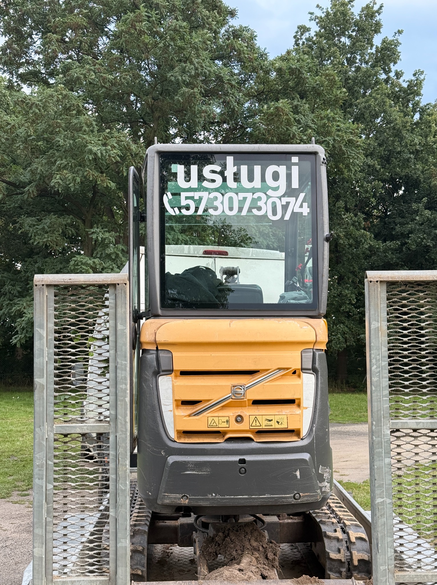Żółto-szara minikoparka Volvo, tyłem, na platformie transportowej; na szybie kabiny naklejka z numerem telefonu 'usługi 573073074', w tle zieleń drzew.