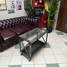 Art-Spaw Goleniów - Metalowy stolik kawowy na kółkach, z siatkowym blatem i dolną półką, stoi na kafelkach. W tle skórzana sofa i dekoracje ścienne.