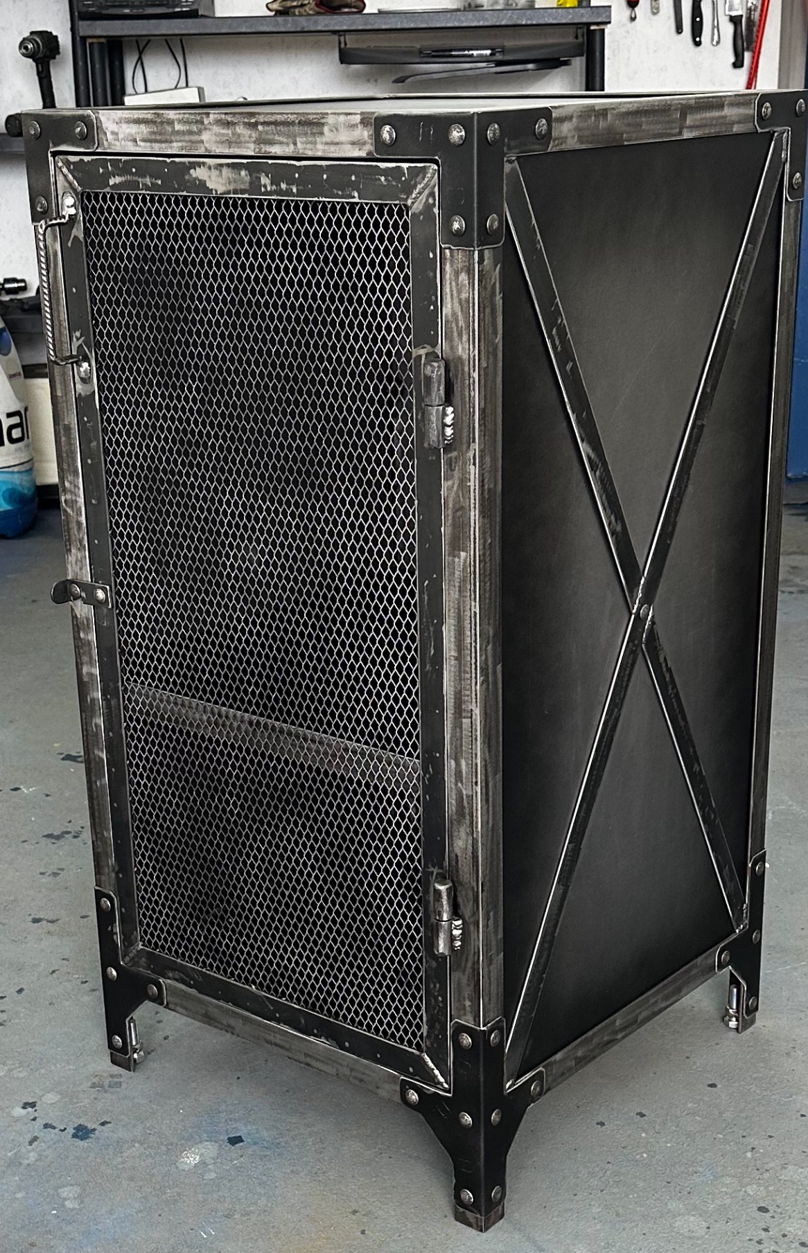 Metalowa komoda w stylu industrialnym z siatkowymi drzwiczkami i dekoracyjnym krzyżem na boku, widoczne nity i ślady ręcznej obróbki metalu.