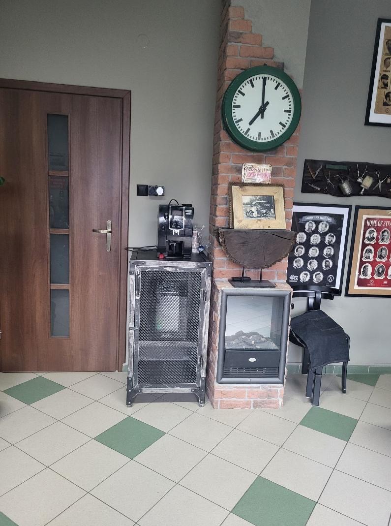 Industrialna komoda z metalową siatką obok ceglanej kolumny z zegarem i elektrycznym kominkiem, na tle wzorzystej podłogi i drewnianych drzwi.