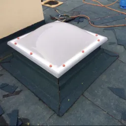 Dach pokryty papą z zamontowanym świetlikiem dachowym. Widoczne narzędzia i butla gazowa. Fragment ściany budynku w tle.