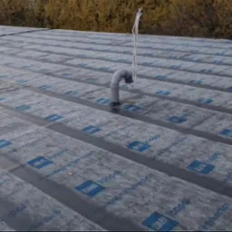 Dach pokryty papą z logo Icopal, z wystającą rurką wentylacyjną. Widoczne pasy papy ułożone równolegle. Tło z drzewami w jesiennej kolorystyce.