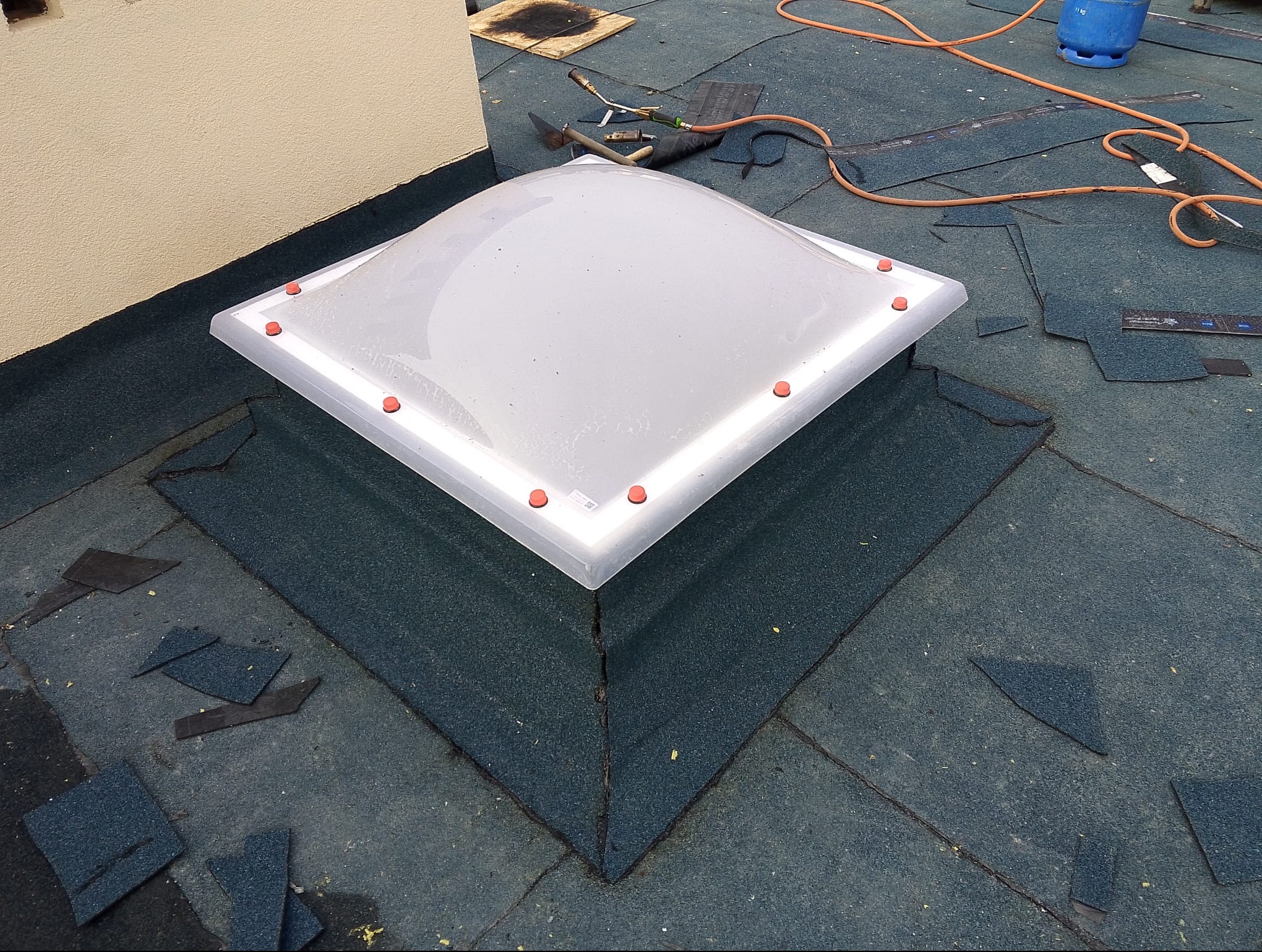 Dach pokryty papą z zamontowanym świetlikiem dachowym. Widoczne narzędzia i butla gazowa. Fragment ściany budynku w tle.