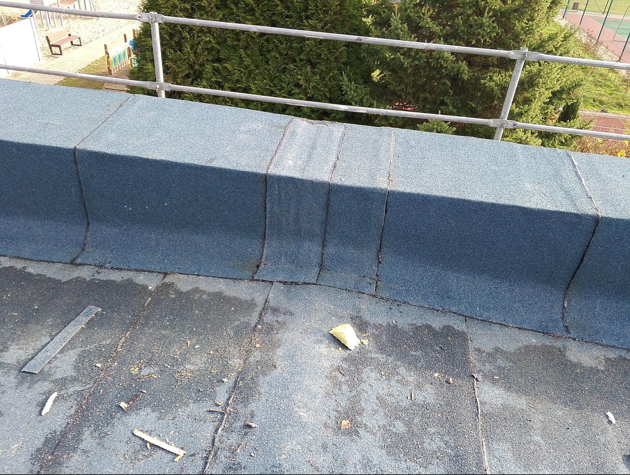 Płaski dach pokryty papą, z widocznymi łączeniami i fragmentami posypki. W tle barierka i zieleń. Detal wykonanej izolacji dachu płaskiego.