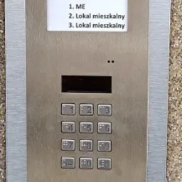 Panel domofonu ACO w metalowej obudowie z klawiaturą numeryczną i wyświetlaczem wyboru lokalu, zamontowany na ścianie o fakturze lastryko.