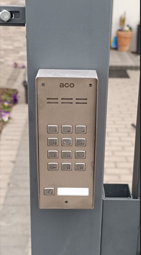 Metalowy panel domofonu ACO z klawiaturą numeryczną, zamontowany na szarym słupku bramy wejściowej. Widoczne śruby mocujące i fragment otoczenia.