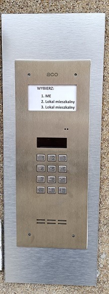 Panel domofonu ACO w metalowej obudowie z klawiaturą numeryczną i wyświetlaczem wyboru lokalu, zamontowany na ścianie o fakturze lastryko.