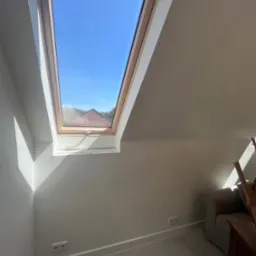Poddasze z oknem dachowym i świeżo pomalowanymi ścianami w odcieniach beżu. Widoczne gniazdka elektryczne i fragment sofy. W tle błękitne niebo i fragment dachu.