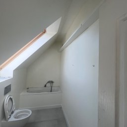 Stepan Struk - Biała łazienka na poddaszu z oknem dachowym, widok na wannę, toaletę i szarą podłogę. Ściany przygotowane pod malowanie, widoczne haczyki na ścianie.