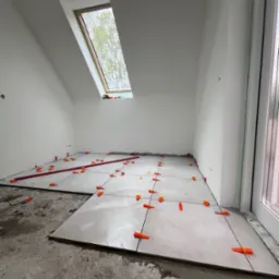 Układanie jasnych płytek podłogowych w pokoju na poddaszu, z oknem dachowym. Widoczne pomarańczowe klipsy poziomujące i czerwona listwa. Etap prac w trakcie realizacji.