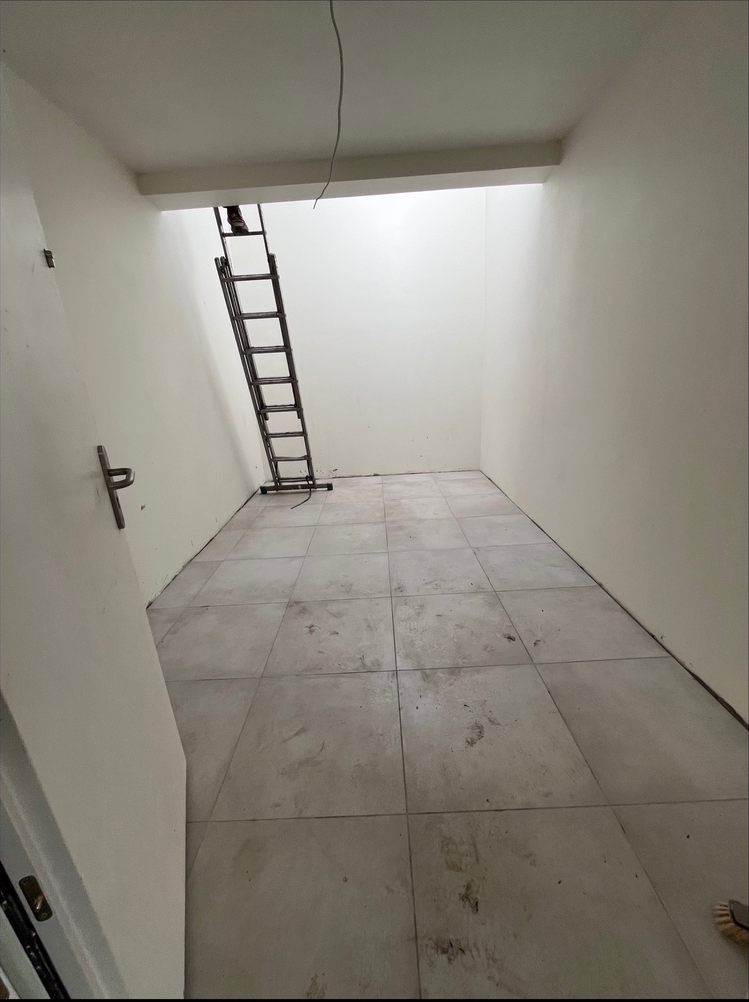 Wnętrze po remoncie: białe ściany, szara podłoga z płytek. Metalowa drabina oparta o ścianę, widoczne ślady po pracy. Minimalistyczny, surowy styl.