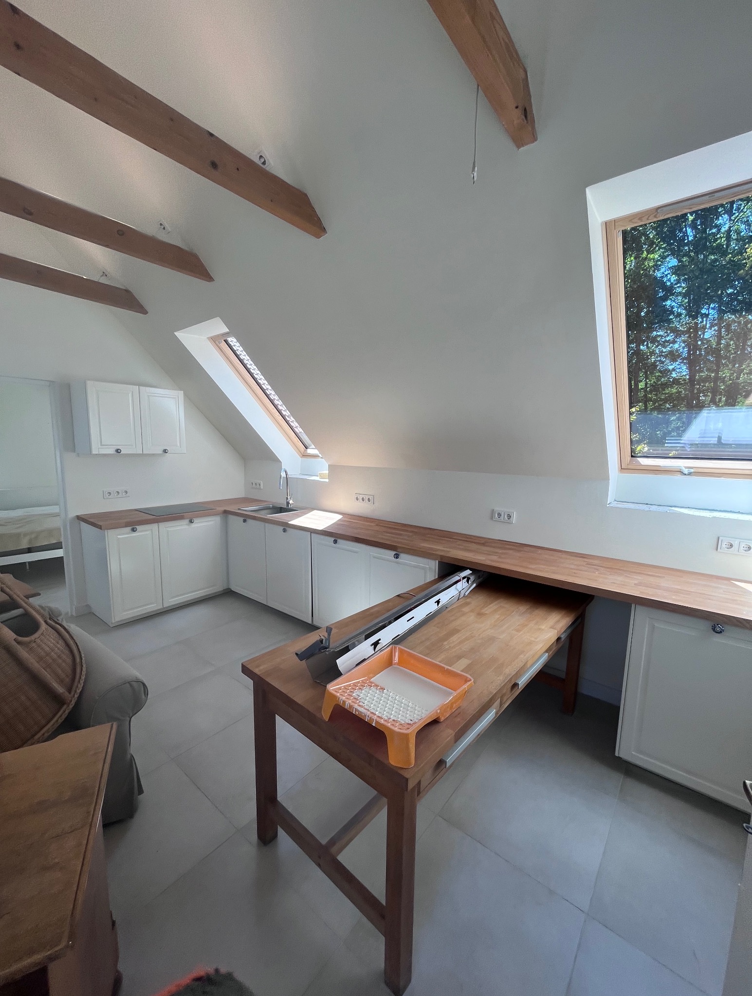 Poddasze z oknem dachowym, białe szafki kuchenne z drewnianym blatem, stół z kuwetą malarską, przygotowany do malowania ścian w jasnym odcieniu.