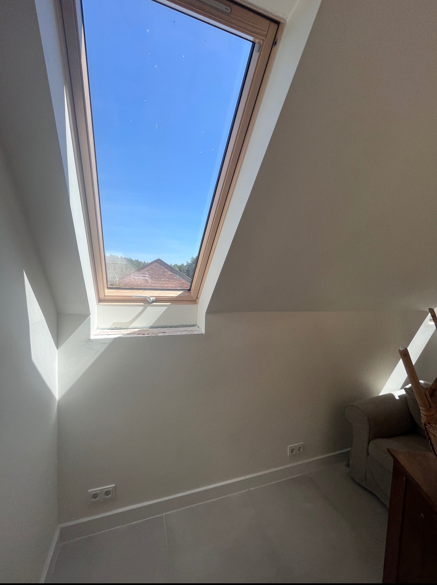 Poddasze z oknem dachowym i świeżo pomalowanymi ścianami w odcieniach beżu. Widoczne gniazdka elektryczne i fragment sofy. W tle błękitne niebo i fragment dachu.