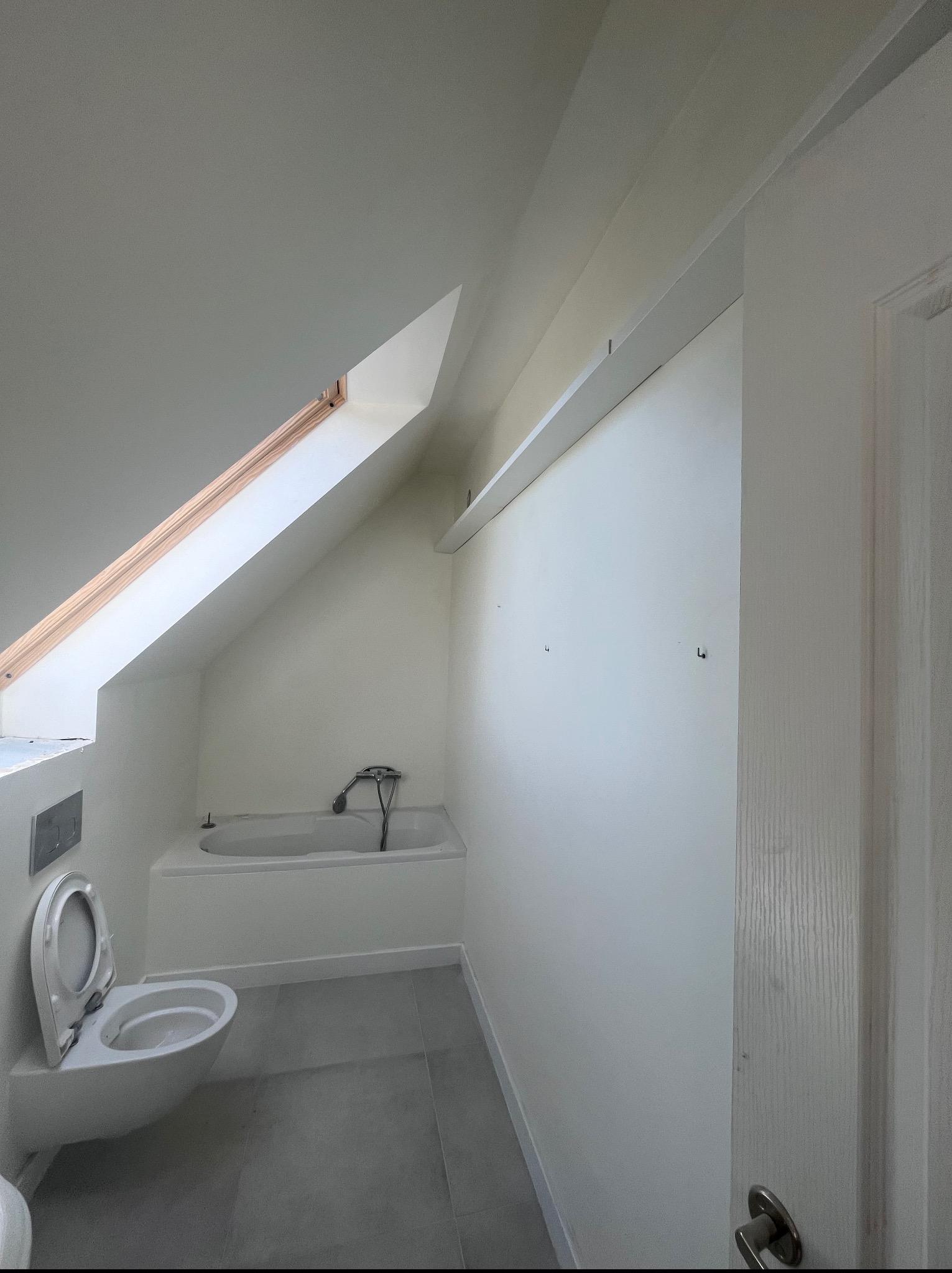 Biała łazienka na poddaszu z oknem dachowym, widok na wannę, toaletę i szarą podłogę. Ściany przygotowane pod malowanie, widoczne haczyki na ścianie.