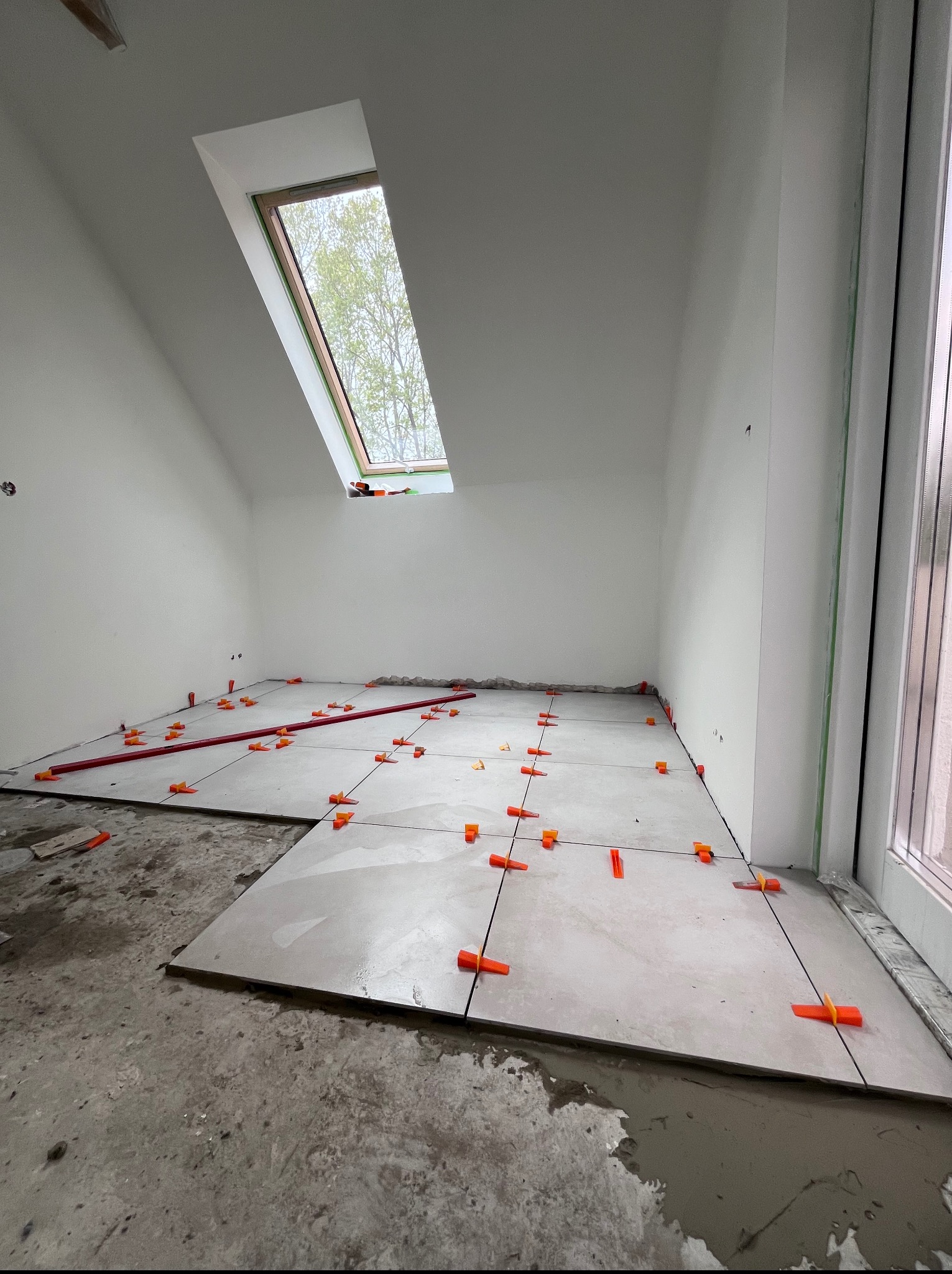 Układanie jasnych płytek podłogowych w pokoju na poddaszu, z oknem dachowym. Widoczne pomarańczowe klipsy poziomujące i czerwona listwa. Etap prac w trakcie realizacji.