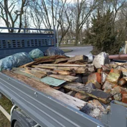 Przyczepa wypełniona gruzem, cegłami i drewnem, z workami odpadów w tle, na tle parku miejskiego w słoneczny dzień. Widoczne elementy konstrukcyjne przyczepy.