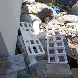 Jakub z Człuchowa - Styropianowe formy, płytki marmurowe i worki z odpadami budowlanymi składowane przy białej ścianie, z fragmentem samochodu w tle, w jasnym oświetleniu.