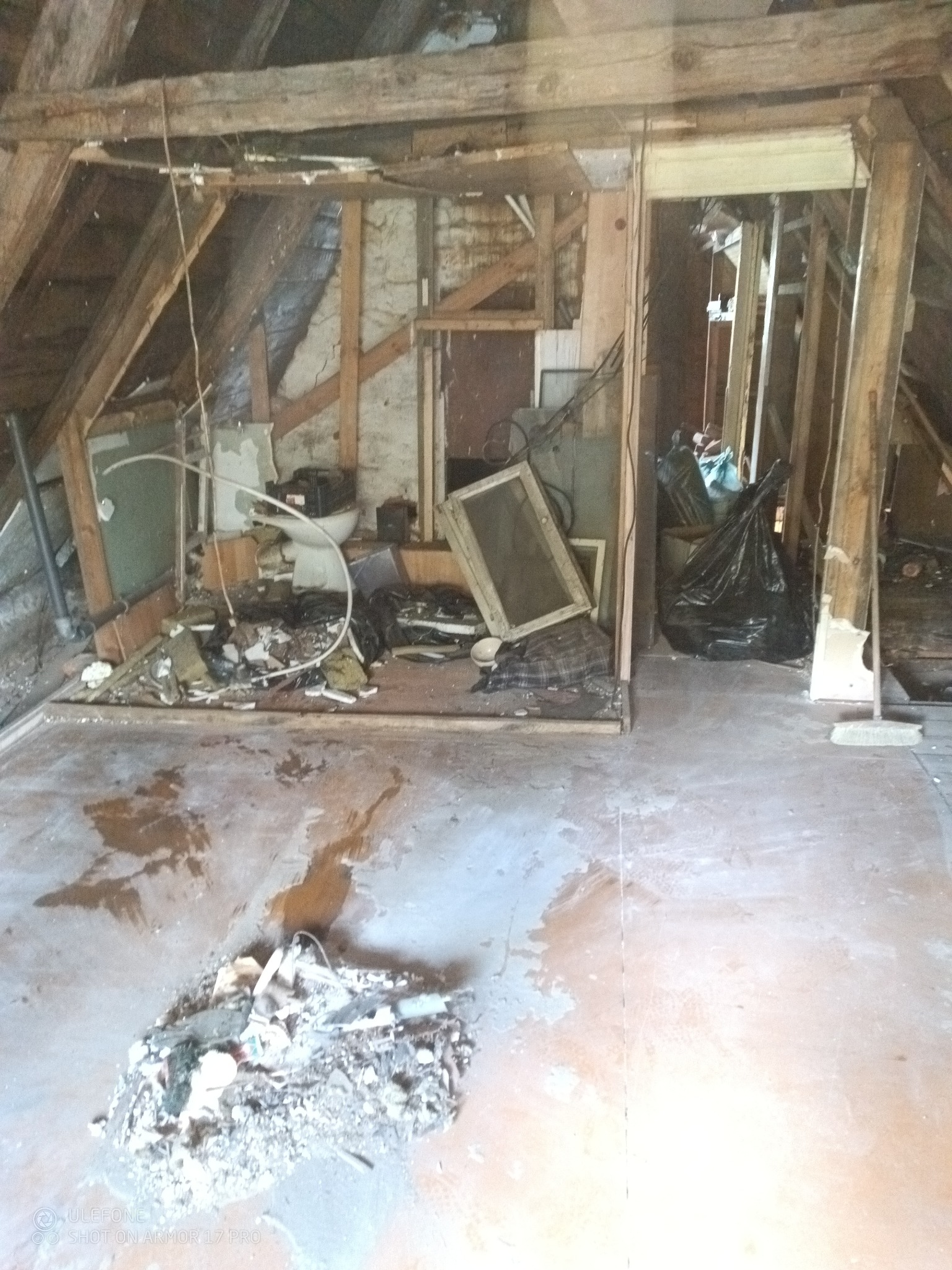 Zdemolowane poddasze ze stertą śmieci, gruzu i odpadów budowlanych. Widoczne elementy konstrukcji dachu i worki na śmieci. Podłoga brudna, z widocznymi plamami.