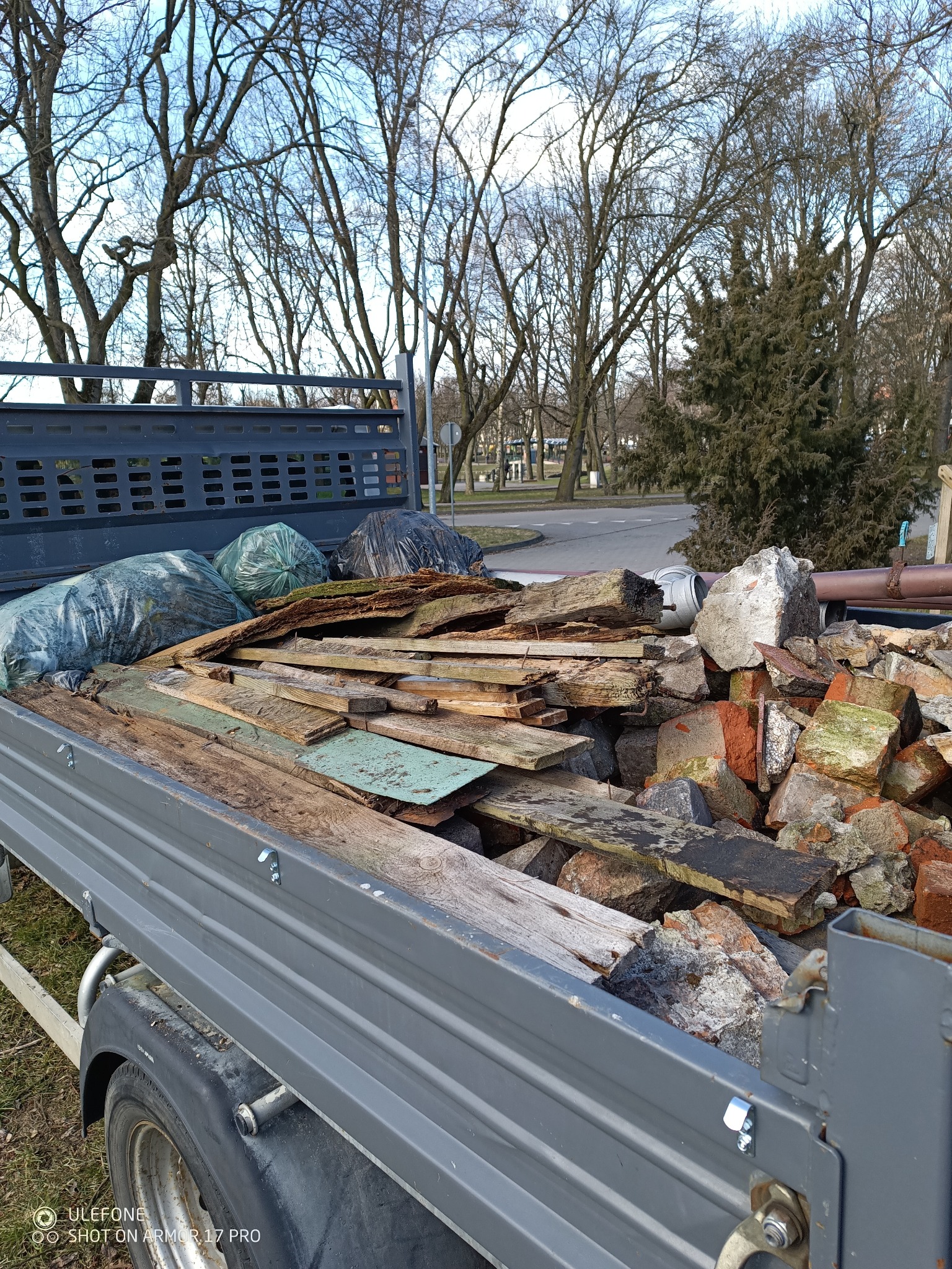 Przyczepa wypełniona gruzem, cegłami i drewnem, z workami odpadów w tle, na tle parku miejskiego w słoneczny dzień. Widoczne elementy konstrukcyjne przyczepy.