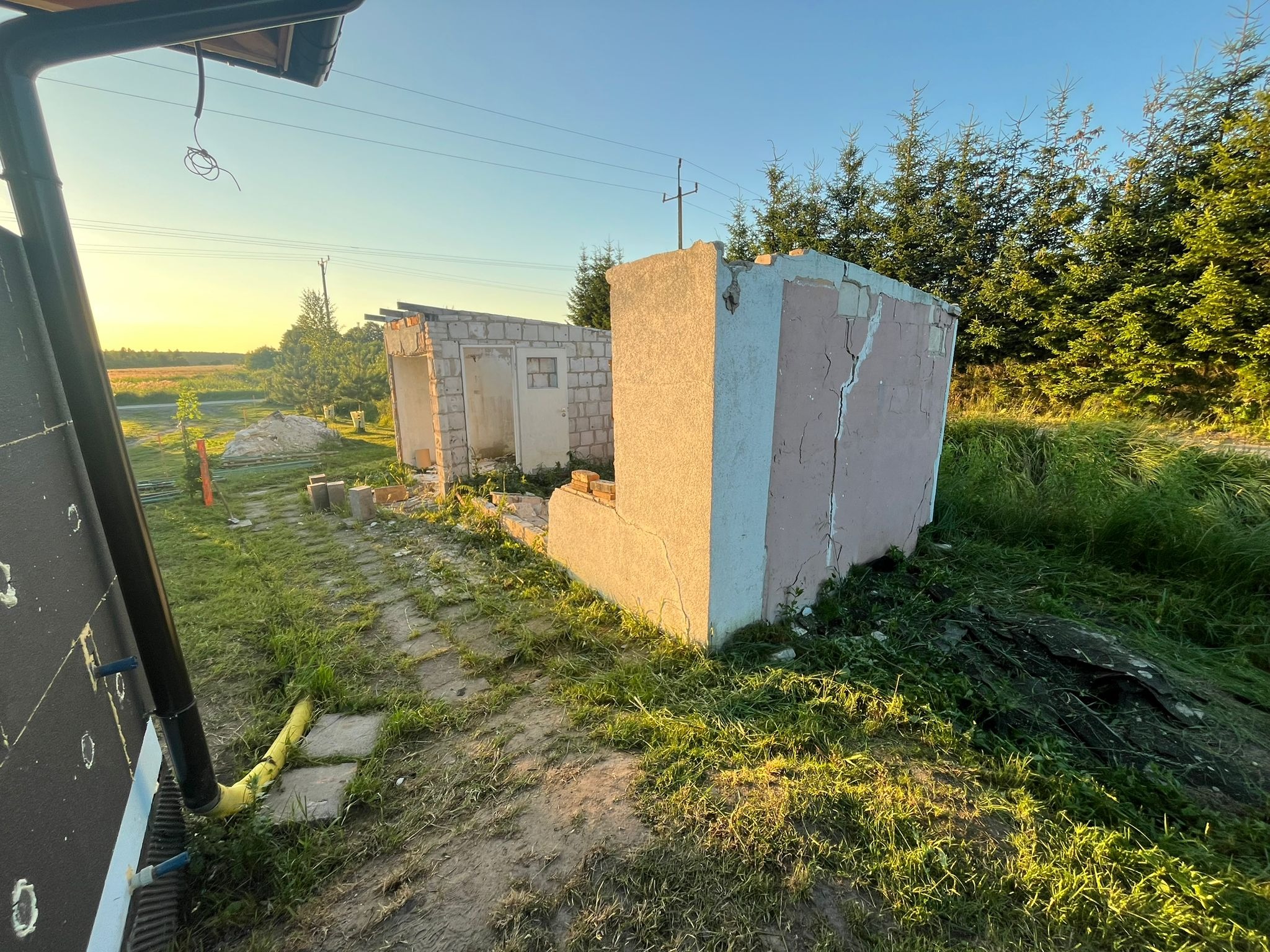 Częściowo rozebrany budynek gospodarczy z cegły i betonu na zarośniętej działce, w tle drzewa i zachodzące słońce. Widoczne ślady rozbiórki.