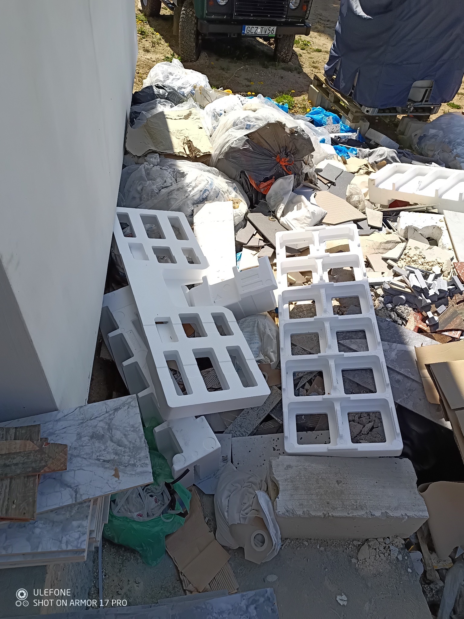 Styropianowe formy, płytki marmurowe i worki z odpadami budowlanymi składowane przy białej ścianie, z fragmentem samochodu w tle, w jasnym oświetleniu.