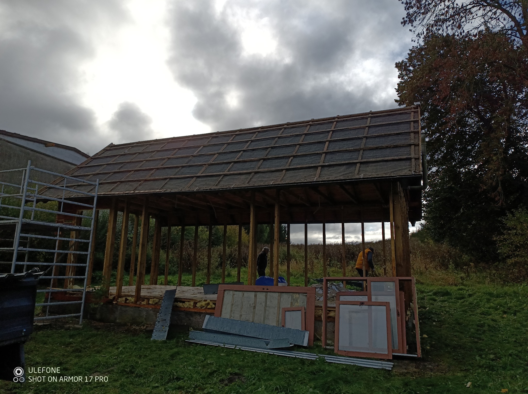 Konstrukcja zadaszenia w trakcie budowy, widoczne drewniane elementy i częściowo ułożone pokrycie. Na pierwszym planie okna i blacha. Prace budowlane w toku.