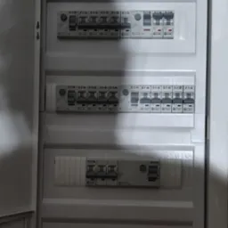 Otwarta, biała skrzynka elektryczna z bezpiecznikami Eaton i modułami Vcx. Widoczne przewody w górnej części. Ściana w tle surowa, z widocznymi śladami po kuciu.