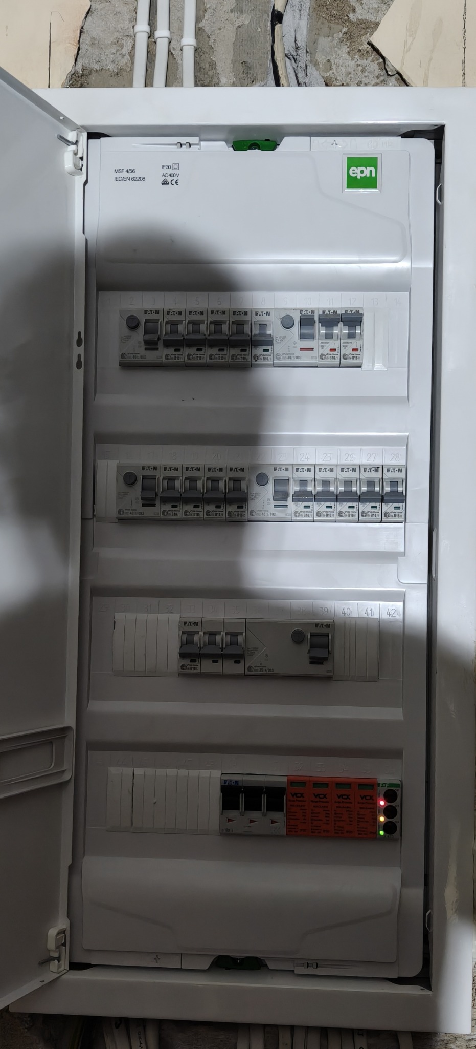 Otwarta, biała skrzynka elektryczna z bezpiecznikami Eaton i modułami Vcx. Widoczne przewody w górnej części. Ściana w tle surowa, z widocznymi śladami po kuciu.