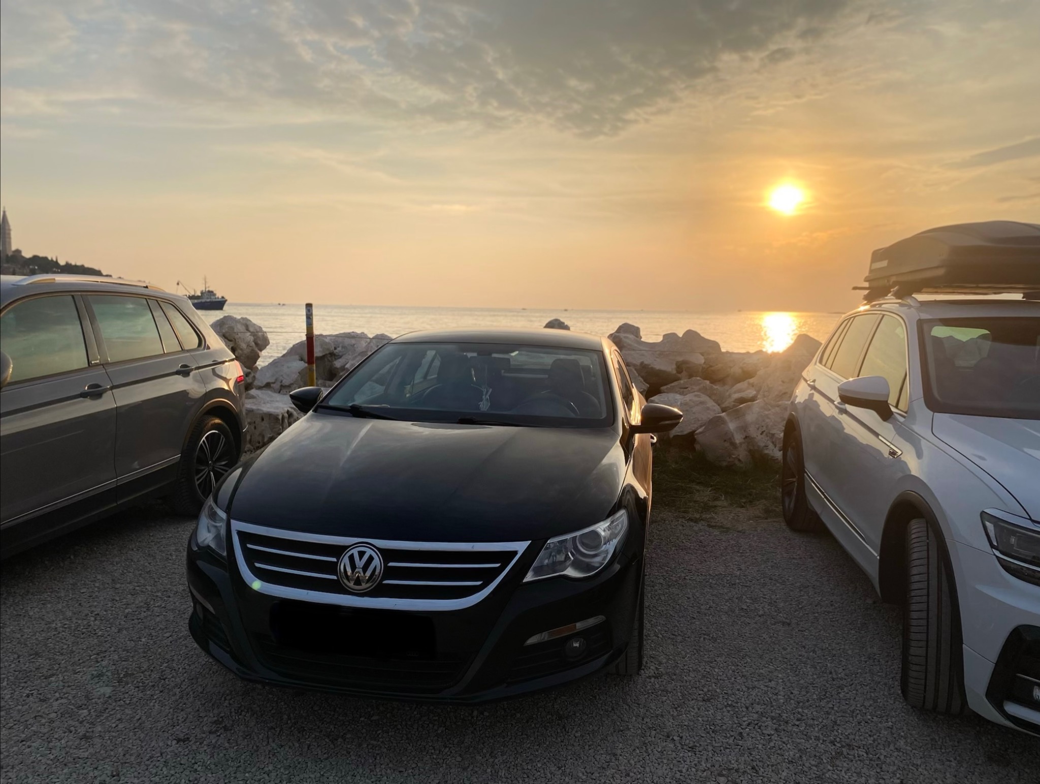Czarny Volkswagen CC na tle zachodzącego słońca nad morzem, obok zaparkowane inne auta. Widok na kamieniste wybrzeże i spokojną wodę.