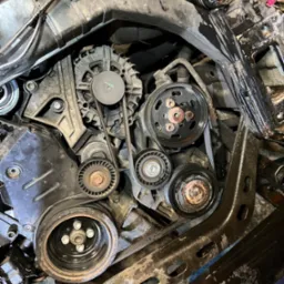 Detaliczny widok wnętrza silnika samochodowego z paskiem klinowym, kołami pasowymi i elementami metalowymi, ukazujący złożoność i stan techniczny.