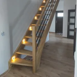 Drewniane schody z oświetleniem LED na stopniach i czarną, metalową balustradą. Widok z dołu na górę, drzwi w tle.