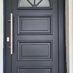 HOLZMET SPÓŁKA CYWILNA SŁAWOMIR MAŚLANY, MATEUSZ MAŚLANY - Ciemne drzwi zewnętrzne z szybą w kształcie półkola i długim, srebrnym uchwytem, osadzone w białej ścianie. Widoczna wycieraczka pod drzwiami.