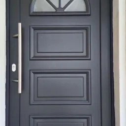 Ciemne drzwi zewnętrzne z szybą w kształcie półkola i długim, srebrnym uchwytem, osadzone w białej ścianie. Widoczna wycieraczka pod drzwiami.