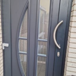 HOLZMET SPÓŁKA CYWILNA SŁAWOMIR MAŚLANY, MATEUSZ MAŚLANY - Grafitowe drzwi wejściowe z szybami o nietypowym kształcie, srebrna klamka, widoczna poziomica przy murze z jasnej cegły. Nowoczesny design, detale wykończenia.