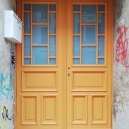 HOLZMET SPÓŁKA CYWILNA SŁAWOMIR MAŚLANY, MATEUSZ MAŚLANY - Dwuskrzydłowe drzwi zewnętrzne w odcieniu miodowym z przeszklonymi górnymi panelami, zamontowane w ścianie z graffiti i skrzynką pocztową.