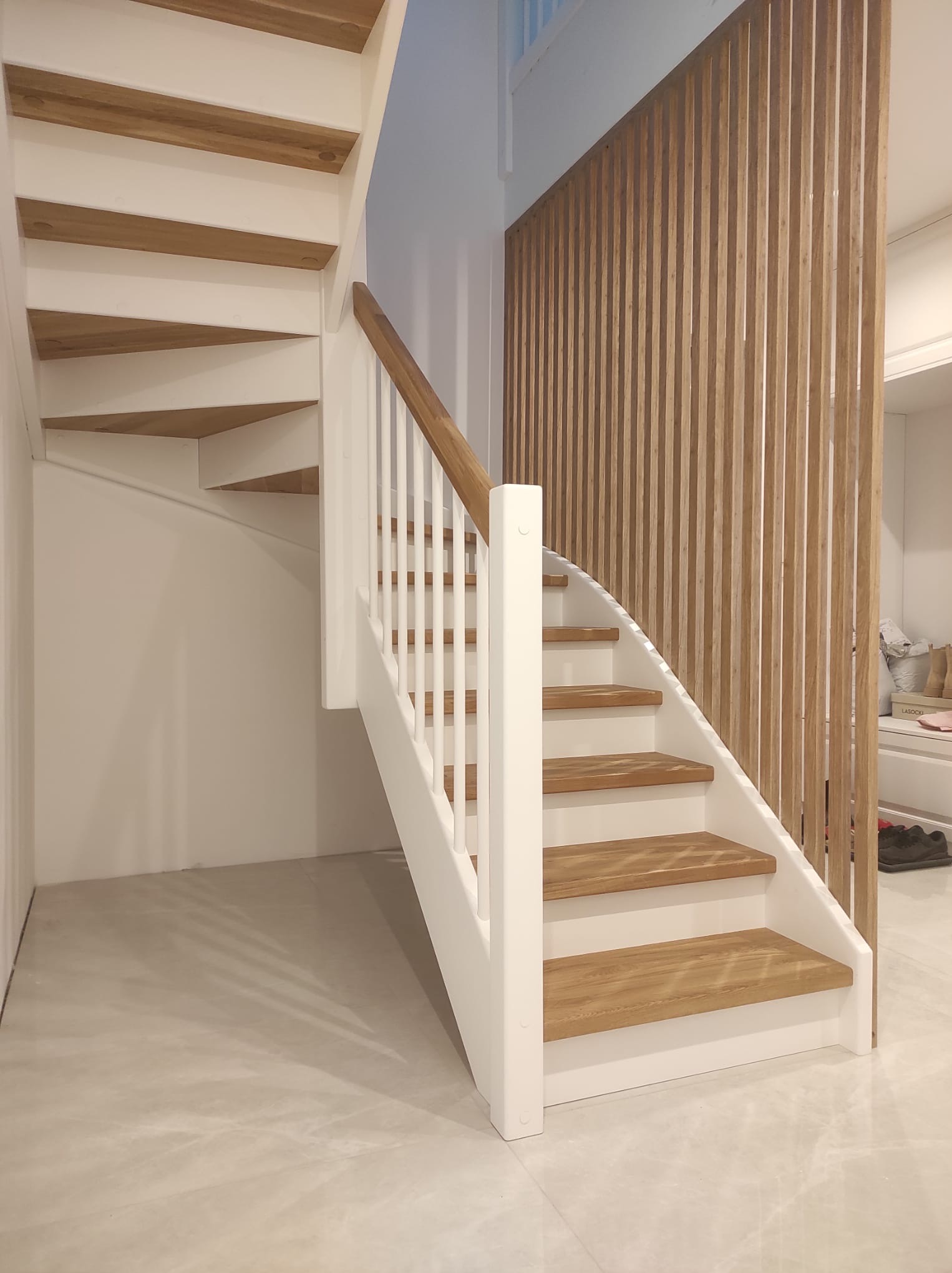 Białe schody z drewnianymi stopniami i balustradą, z dekoracyjną drewnianą ścianką działową w tle. Widok z dołu, podkreślający zakrzywiony kształt schodów.