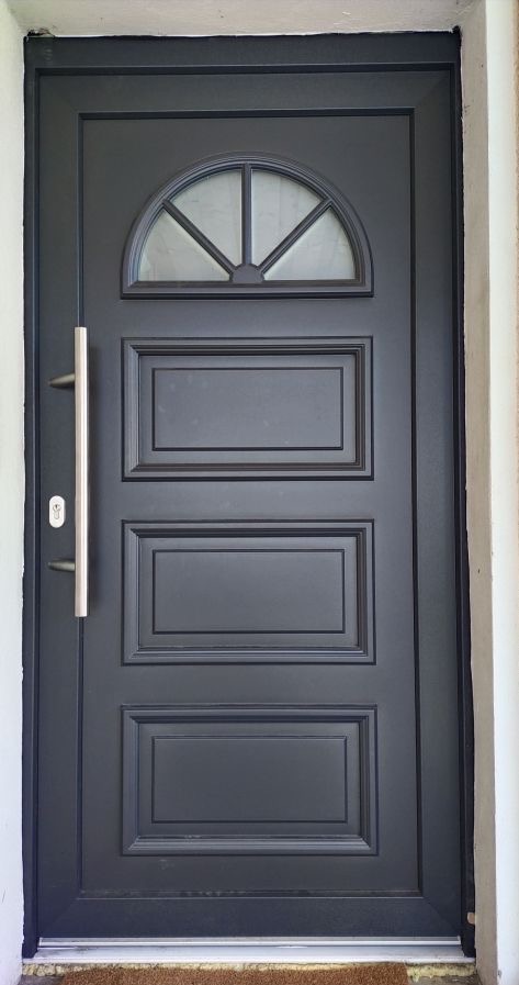 Ciemne drzwi zewnętrzne z szybą w kształcie półkola i długim, srebrnym uchwytem, osadzone w białej ścianie. Widoczna wycieraczka pod drzwiami.