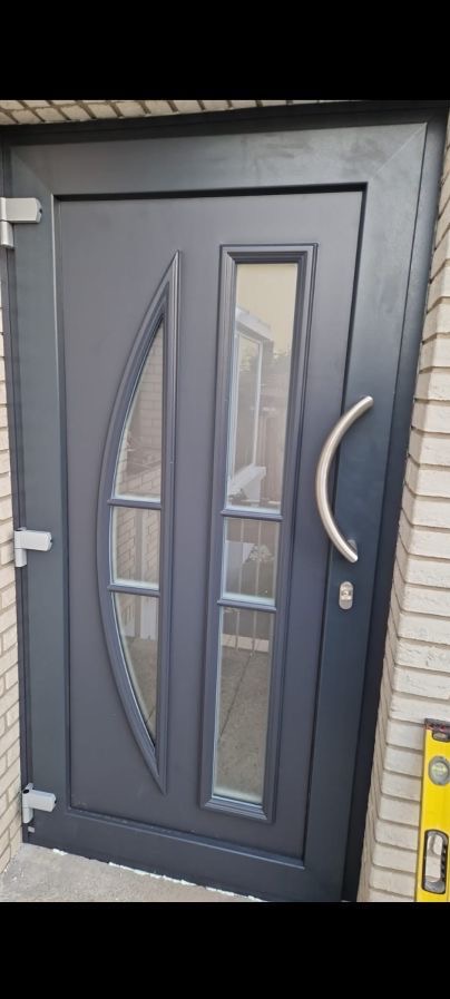 Grafitowe drzwi wejściowe z szybami o nietypowym kształcie, srebrna klamka, widoczna poziomica przy murze z jasnej cegły. Nowoczesny design, detale wykończenia.