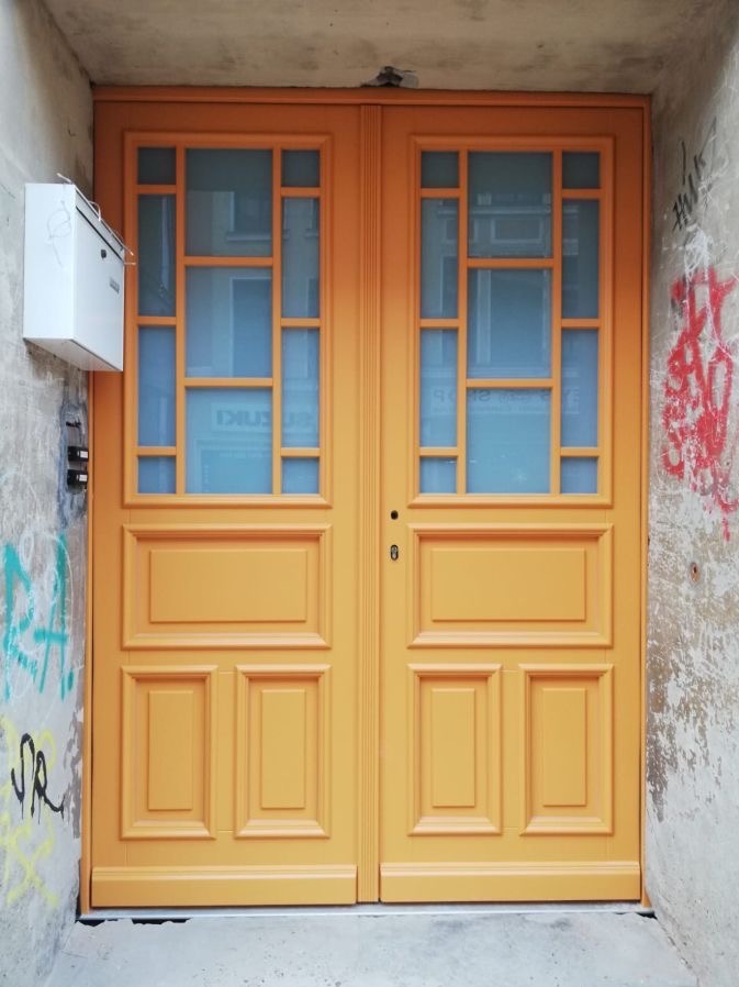 Dwuskrzydłowe drzwi zewnętrzne w odcieniu miodowym z przeszklonymi górnymi panelami, zamontowane w ścianie z graffiti i skrzynką pocztową.