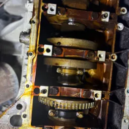 Otwarty silnik samochodowy z widocznymi elementami wewnętrznymi, takimi jak wał korbowy i zębatka, pokryte olejem. Widoczne ślady użytkowania i zużycia.