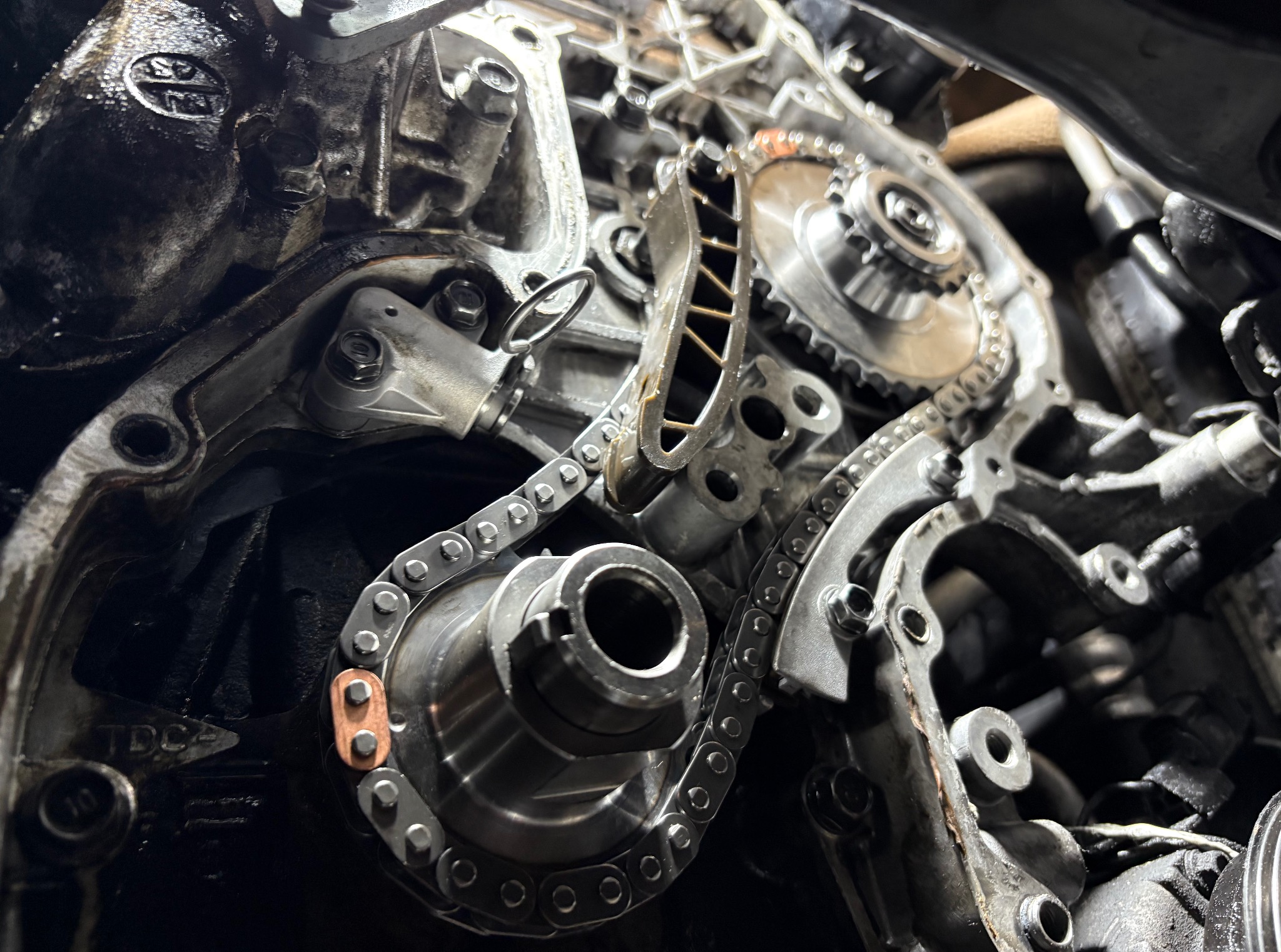 Otwarty silnik samochodowy z widocznym łańcuchem rozrządu i zębatkami. Elementy metalowe, ślady oleju. Detal mechanizmu w trakcie naprawy lub serwisu.