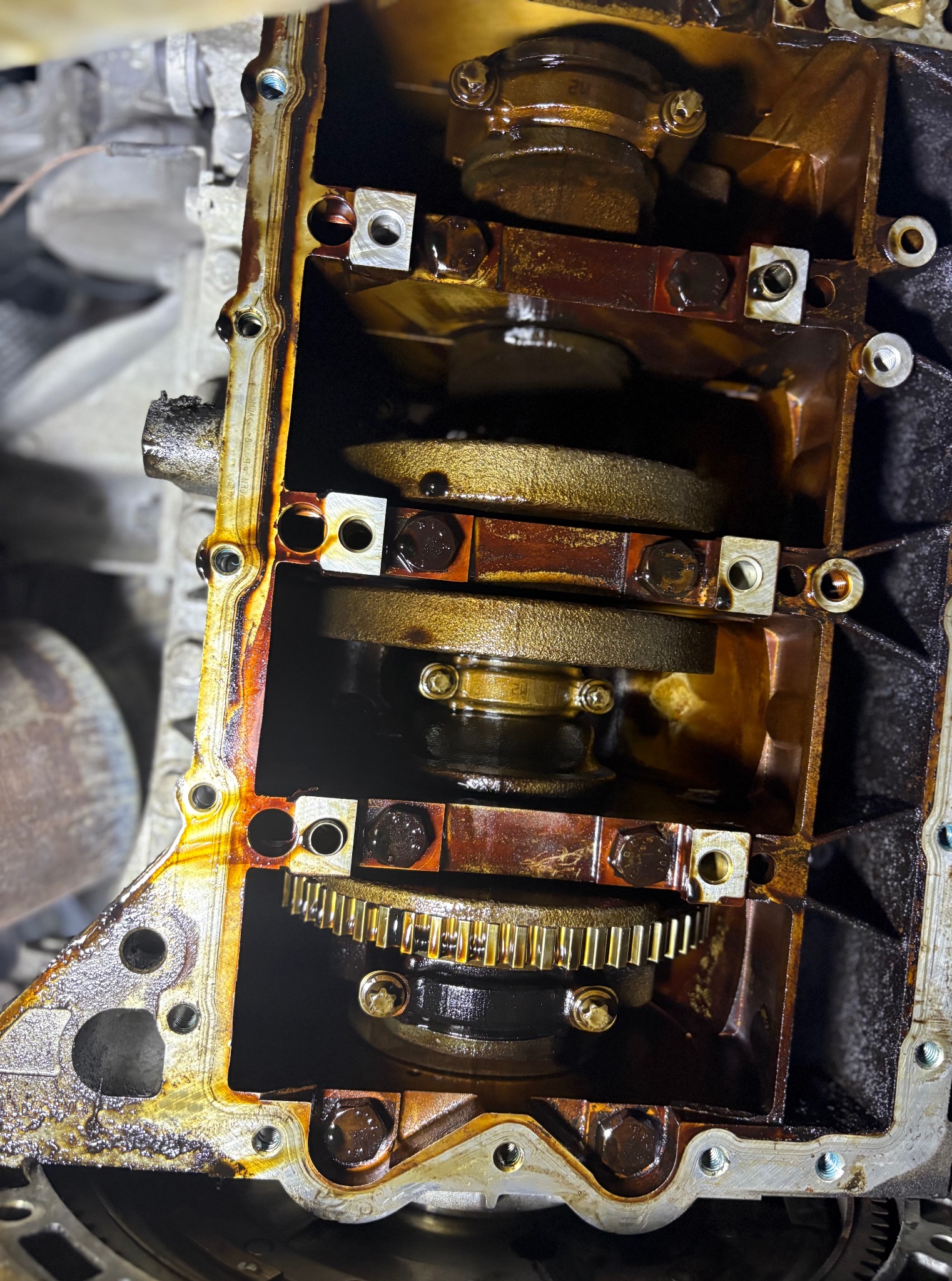 Otwarty silnik samochodowy z widocznymi elementami wewnętrznymi, takimi jak wał korbowy i zębatka, pokryte olejem. Widoczne ślady użytkowania i zużycia.