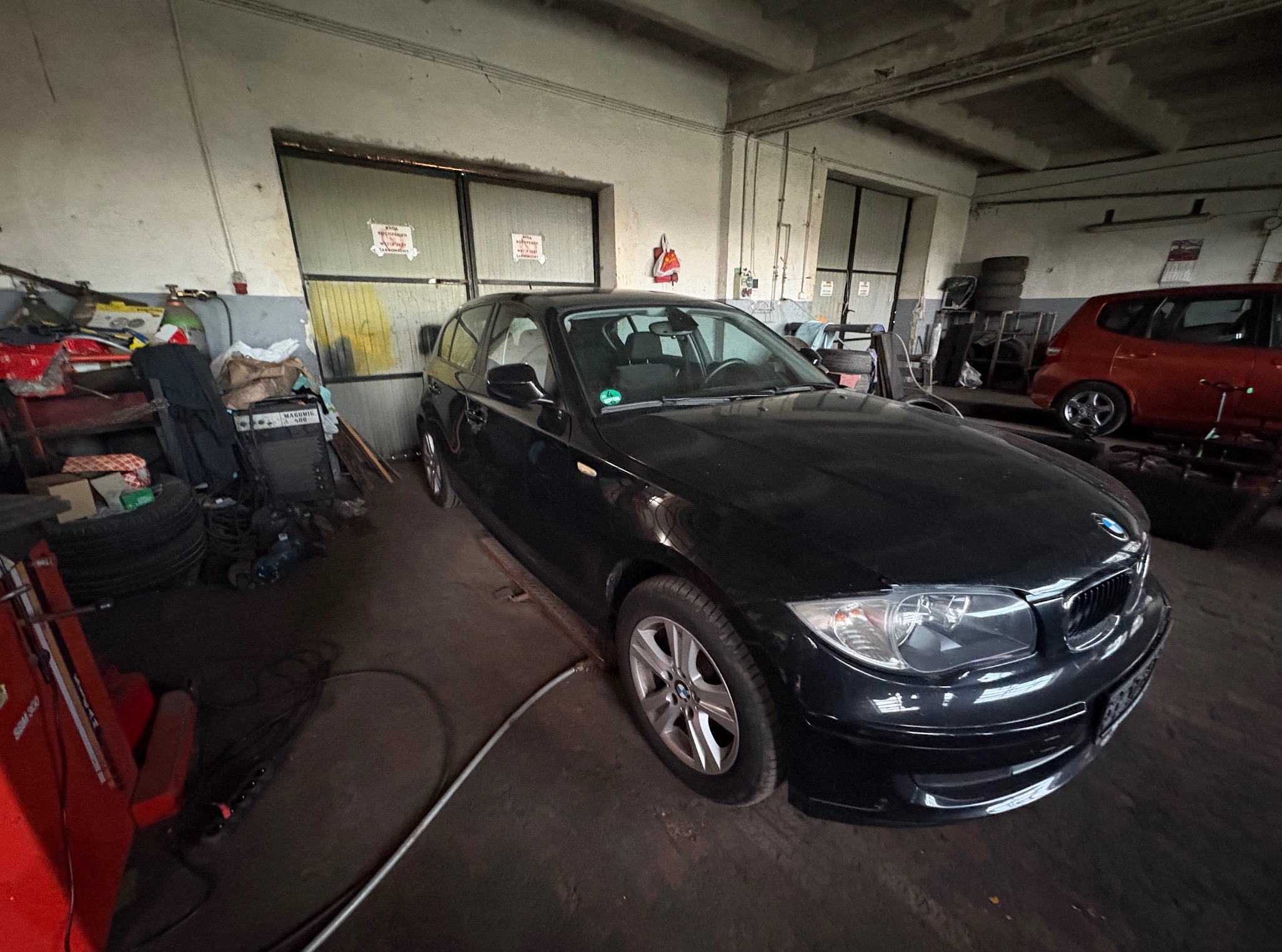 Ciemny BMW serii 1 w warsztacie samochodowym; widoczne narzędzia i wyposażenie warsztatu, w tle czerwony samochód.