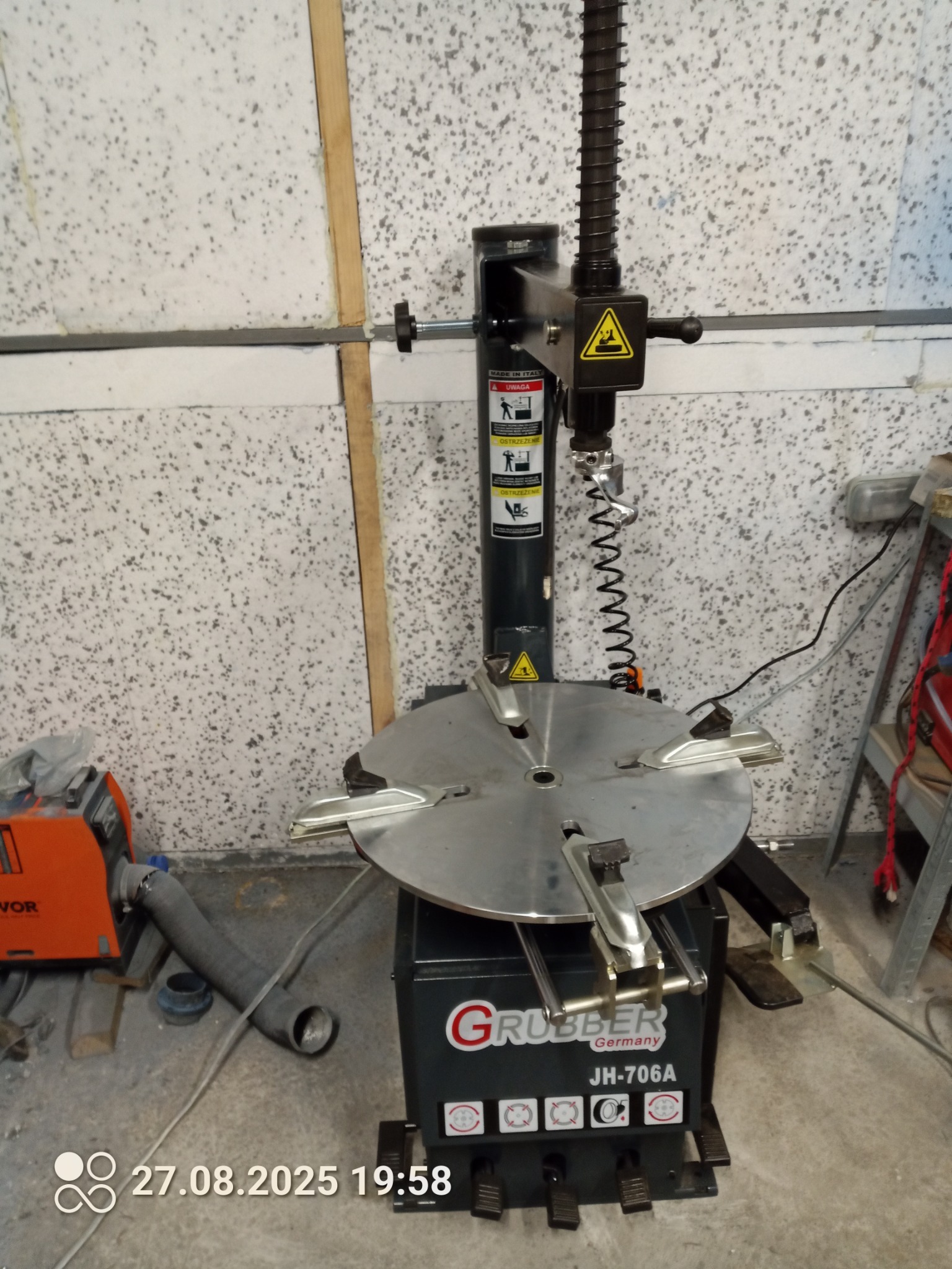 Montażownica do opon Gruber JH-706A w warsztacie, z widocznymi elementami sterującymi i oznaczeniami ostrzegawczymi. Tło stanowi ściana z fakturą.