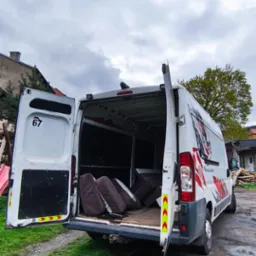 Otwarty biały bus z numerem 67 i grafiką, załadowany brązowymi poduszkami na trawiastym podwórku pod pochmurnym niebem. Widoczne otwarte drzwi i wnętrze pojazdu.