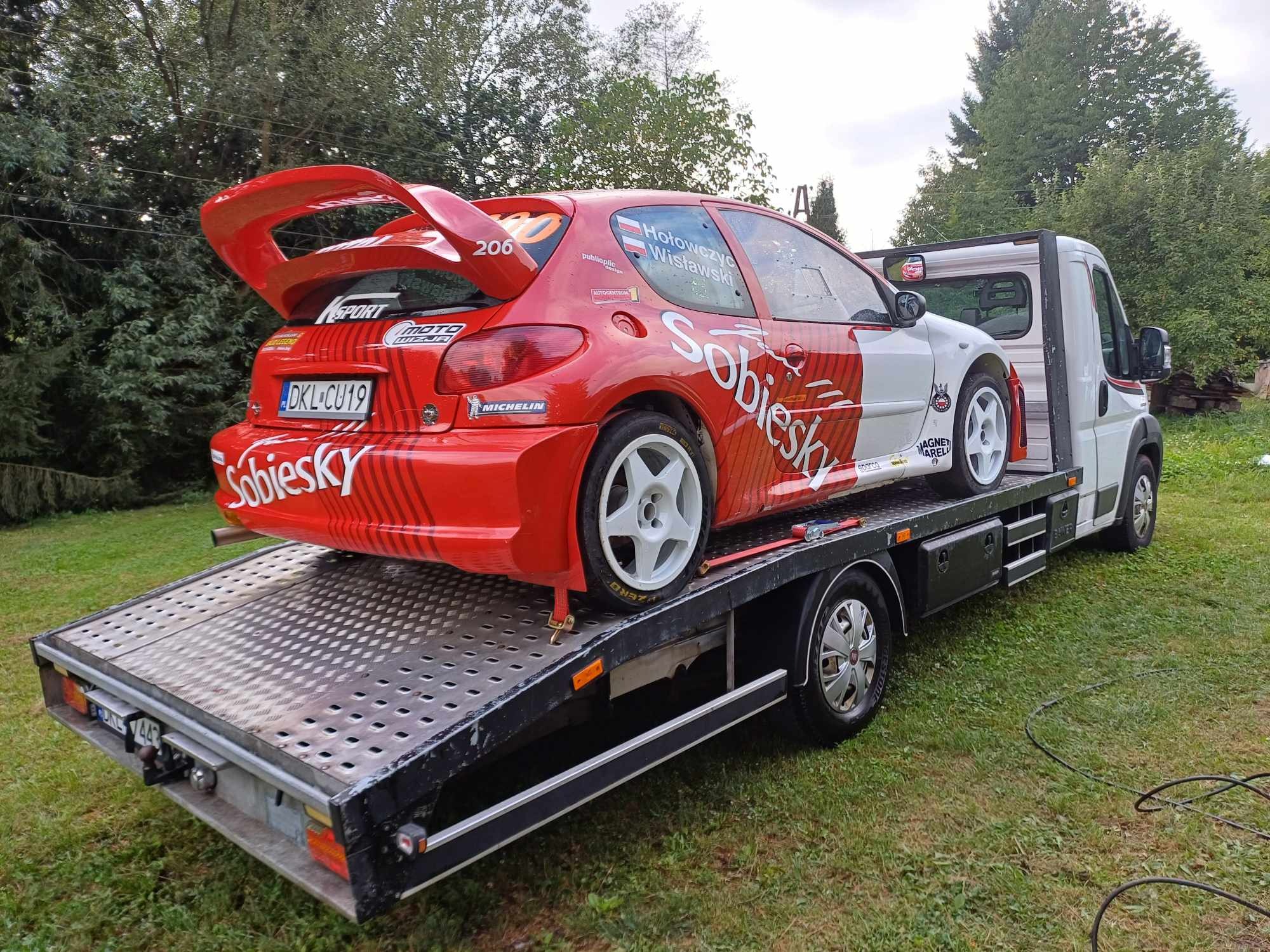 Czerwono-biały Peugeot 206 rajdowy z dużym spojlerem na lawecie. Widoczne detale: logo Sobiesky, numer 206, tablica DKL CU19, zieleń w tle.