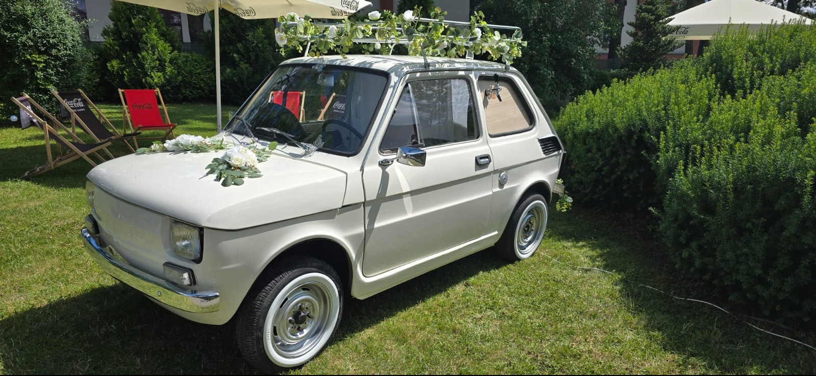 Biały, udekorowany kwiatami Fiat 126p na trawie, z elementami dekoracji ślubnych. W tle krzesła plażowe i parasole Coca-Cola. Nietypowy pojazd na uroczystości.