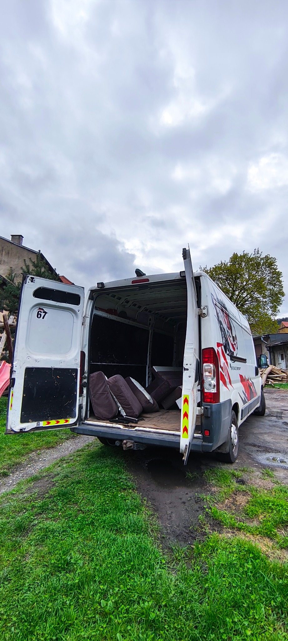 Otwarty biały bus z numerem 67 i grafiką, załadowany brązowymi poduszkami na trawiastym podwórku pod pochmurnym niebem. Widoczne otwarte drzwi i wnętrze pojazdu.
